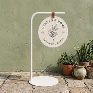 Op de afbeelding: Een witte metalen standaard met een ronde voet en een hangend rond bord. Het bord is crèmekleurig met een zwarte botanische illustratie en de tekst "GARDEN & HOME BOTANICAL WARES EST. 2023". Het bord is bevestigd met een bruine leren riem.