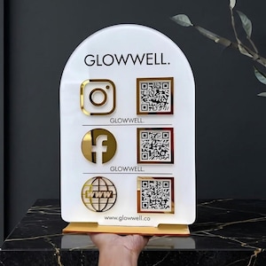 Könnte beinhalten: Ein bogenförmiges weißes Acrylschild mit dem schwarzen Text GLOWWELL. oben und www.glowwell.co unten. Es zeigt drei goldene Social-Media-Symbole auf der linken Seite und drei entsprechende goldene QR-Codes auf der rechten Seite.