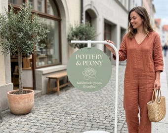 Puede incluir: Un soporte de letrero de metal blanco con un letrero redondo de color verde salvia que presenta el texto blanco POTTERY & PEONY, un dibujo lineal de una flor de peonía y el texto handmade goods & coffee, colocado en una calle adoquinada.