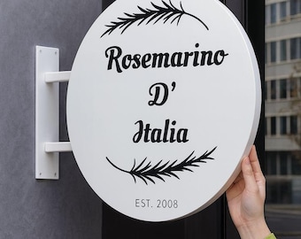 Puede incluir: Un letrero redondo blanco con el texto "Rosemarino D' Italia" en escritura negra, con ramitas decorativas. El letrero está montado sobre un soporte blanco y dice "EST. 2008".