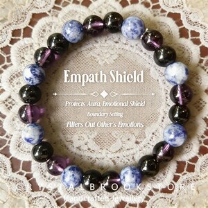 May include: A bracelet with the text "Empath Shield" made of black, purple, and blue-grey spherical beads. The image text says it protects the aura, sets boundaries, and filters emotions. Handcrafted jewellery.