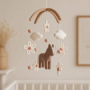 Könnte beinhalten: Ein Baby-Mobile mit einem Holzbogen, an dem verschiedene Elemente hängen. Es zeigt ein gestricktes braunes Pferd, weiße Wolken, cremefarbene Blumen mit rosa Zentren und rosa Filzkugeln. Für das Kinderzimmer.