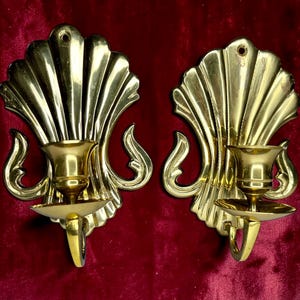 May include: A pair of polished brass wall sconces, each featuring a large, fan-shaped shell backplate with vertical ridges and a central candle holder cup. The sconces have a curved, decorative base support and a small hole at the top for mounting.