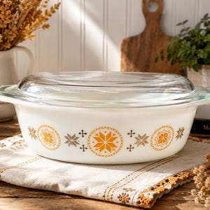 May include: A white casserole dish with a clear glass lid. The dish features a decorative pattern of orange and brown starburst and floral designs. The dish is sitting on a cream-colored cloth with an orange trim.