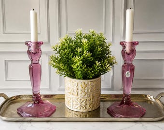 May include: Two translucent pink glass candle holders with white taper candles, a small potted green plant in a white and gold patterned ceramic pot, and a rectangular metallic silver tray with two handles on the sides.