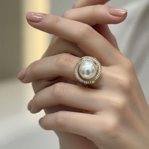 May include: A gold ring featuring a large, round, white pearl at the center, encircled by a halo of small, clear gemstones. The ring's band has a twisted design. The ring is on a person's finger.