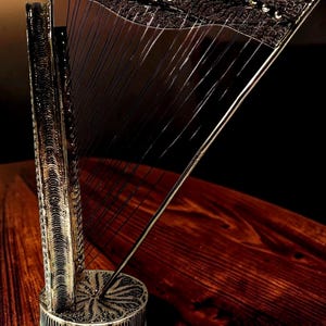 May include: A decorative miniature harp with a dark, textured finish. The harp features intricate detailing on the soundbox and neck, with strings stretched across the frame. The harp is set on a wooden surface, showcasing its elegant design.