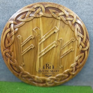 Op de afbeelding: Een ronde houten plaat met een uitgesneden rand van ingewikkelde Keltische knoopmotieven. Het midden toont drie verhoogde runensymbolen. De tekst RUSTIC MART HANDCRAFTED is in het zwart gedrukt onderaan het midden van het lichtbruine hout.