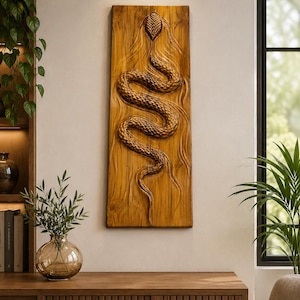May include: A rectangular wooden wall plaque featuring a detailed, three-dimensional relief carving of a snake winding vertically across the centre. The natural wood grain is visible, with the snake's scales textured and defined in a warm brown tone.