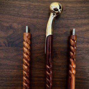 May include: A walking stick assembly consisting of three distinct segments. Two segments feature a dark brown, spiraled wooden shaft with a threaded metal bolt at the top. The third segment includes a polished gold-tone metal handle.