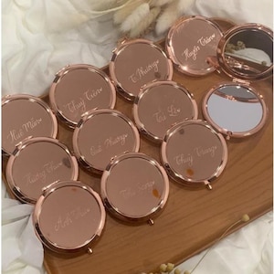 May include: A collection of rose gold compact mirrors, each with a personalized name engraved in a delicate script. One mirror is open, revealing a reflective surface. The mirrors are arranged on a wooden tray, creating a cohesive display.