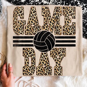 Könnte beinhalten: Ein beiges T-Shirt mit dem Schriftzug GAME DAY in großen Blockbuchstaben mit braun-schwarzem Leopardenmuster. Eine schwarze Volleyball-Grafik ist zwischen den Wörtern zentriert und von drei horizontalen schwarzen Linien flankiert.
