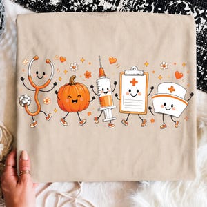 May include: A beige t-shirt featuring a graphic print of five anthropomorphic medical items: an orange stethoscope, a smiling pumpkin, a syringe filled with orange liquid, a clipboard with a medical cross, and a white nurse cap with a cross.