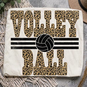 May include: A cream-coloured t-shirt featuring the text VOLLEY BALL in a bold, blocky font with a brown and black leopard print pattern. A black volleyball graphic is centred between the words, flanked by three horizontal black stripes.