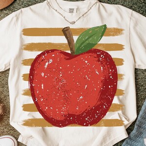 Peut inclure: Un t-shirt à manches courtes de couleur crème arborant un grand motif de pomme rouge vieillie avec une tige brune et une feuille verte. La pomme est centrée sur un fond de rayures horizontales texturées de couleur jaune moutarde.