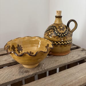 May include: A ceramic cruet with a cork stopper and a matching bowl with a ruffled rim. Both pieces feature a mustard yellow glaze with dark brown hand-painted floral and wavy line patterns. The cruet has a single handle and a rounded body.