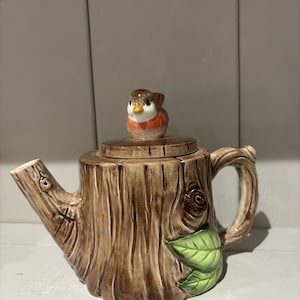 以下が含まれることがあります： 茶色の切り株の形をした陶器製のティーポットで、樹皮のような質感の仕上げが施されています。蓋には赤い胸の小さな鳥のフィギュアが付いています。持ち手と注ぎ口は木の枝を模しており、本体の側面には2枚の緑色の葉が付いています。