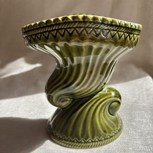 May include: A glossy olive green ceramic vase featuring a flared, scalloped rim and a base with intricate, embossed geometric patterns. The body of the vase displays a swirling, ribbed design that creates a sense of movement and depth in the glaze.