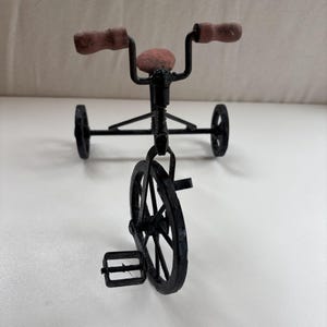 Op de afbeelding: Een miniatuur driewieler van zwart metaal met een voorwiel met pedalen, twee achterwielen en bruine, getextureerde handvatten op het stuur. Het zadel is ook bruin en getextureerd, passend bij de handvatten. Het frame is gemaakt van dunne metalen staven.
