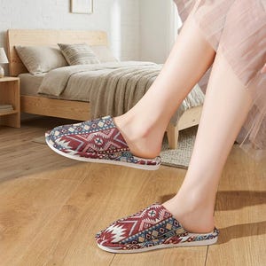 May include: Pair of patterned slippers with a geometric design in red, blue, and cream. The slippers have a slip-on style with a white sole. The image shows the slippers on a wooden floor, suggesting indoor use.