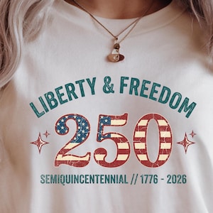 Puede incluir: Camiseta color crema con el texto "Liberty & Freedom 250" en verde azulado y rojo, celebrando el semiquincentenario. El n&uacute;mero 2 y el cero est&aacute;n dise&ntilde;ados con un patr&oacute;n de bandera patri&oacute;tica. La camiseta tambi&eacute;n incluye los a&ntilde;os 1776-2026.