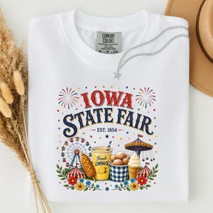 May include: A white t-shirt featuring a colorful graphic with the text "IOWA STATE FAIR EST. 1854". The design includes a Ferris wheel, a corn dog, a cup of fresh lemonade, a bowl of fried dough, a carousel, and fireworks with floral accents.