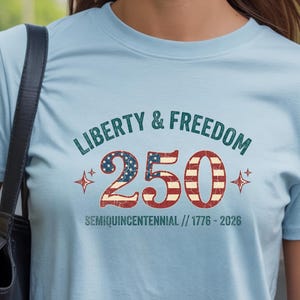 Puede incluir: Camiseta azul claro con las palabras "Liberty & Freedom" sobre el n&uacute;mero "250" en rojo, blanco y azul, con el texto "Semiquincentennial // 1776-2026" debajo. La camiseta tiene cuello redondo y mangas cortas.