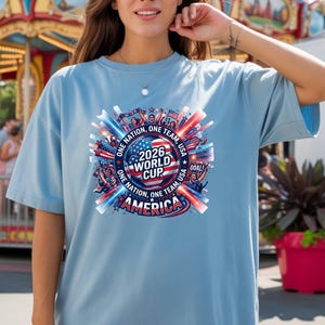 May include: Light blue t-shirt with a circular graphic for the 2026 World Cup in America. The design includes the text "ONE NATION, ONE TEAM, USA" and "AMERICA" with red, white, and blue accents. The shirt is a casual style.
