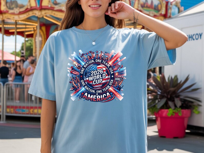 May include: Light blue t-shirt with a circular graphic for the 2026 World Cup in America. The design includes the text "ONE NATION, ONE TEAM, USA" and "AMERICA" with red, white, and blue accents. The shirt is a casual style.