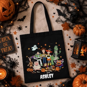 May include: A black tote bag featuring a graphic print of Toy Story characters dressed in Halloween costumes, including a ghost, pumpkins, and the name ASHLEY printed in white text at the bottom of the central illustration.