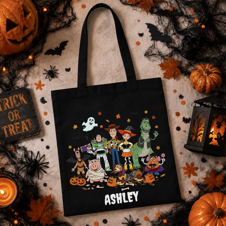 May include: A black tote bag featuring a graphic print of Toy Story characters dressed in Halloween costumes, including a ghost, pumpkins, and the name ASHLEY printed in white text at the bottom of the central illustration.