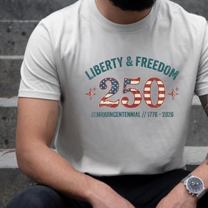 Puede incluir: Camiseta gris claro con el texto "LIBERTY & FREEDOM 250 SEMIQUICENTENNIAL // 1776-2026" en rojo, blanco y azul. El n&uacute;mero 250 est&aacute; dise&ntilde;ado con la bandera americana. La camiseta es de cuello redondo.
