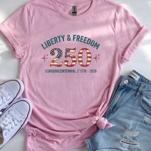 Puede incluir: Una camiseta rosa claro con el texto "LIBERTY & FREEDOM 250 SEMIQUICENTENNIAL // 1776-2026" en un estilo vintage. La camiseta se combina con pantalones cortos de mezclilla y zapatillas blancas.