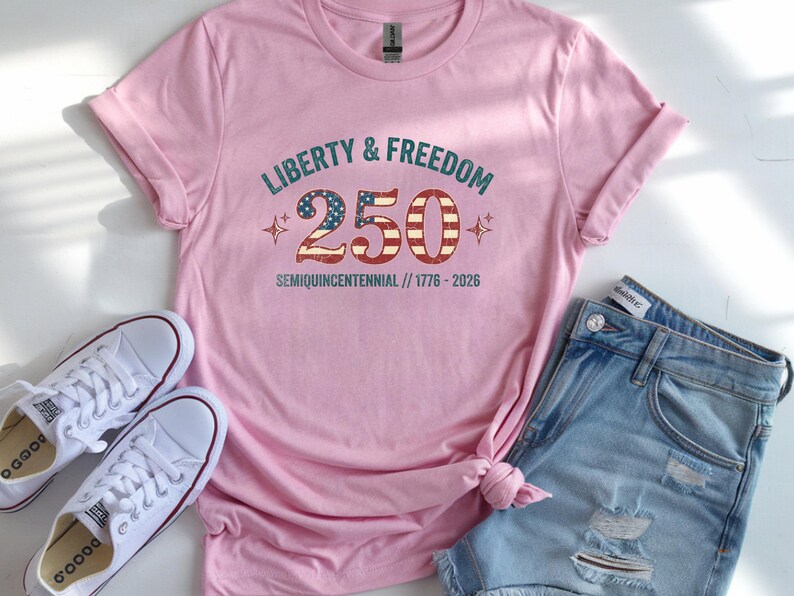 Puede incluir: Una camiseta rosa claro con el texto "LIBERTY & FREEDOM 250 SEMIQUICENTENNIAL // 1776-2026" en un estilo vintage. La camiseta se combina con pantalones cortos de mezclilla y zapatillas blancas.
