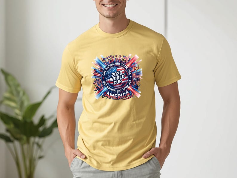 May include: Mustard yellow t-shirt featuring a colorful graphic for the 2026 World Cup in America. The design includes flags, the text "2026 World Cup America", and the phrases "One Nation, One Team, USA".