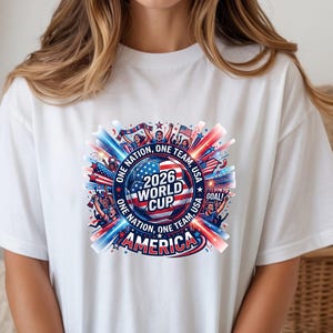 May include: White t-shirt featuring a colorful graphic design for the 2026 World Cup. The design includes a soccer ball, the text "2026 WORLD CUP AMERICA", and the phrases "ONE NATION, ONE TEAM, USA" in red, white, and blue.