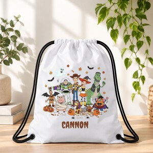 May include: A white drawstring bag featuring a colorful Halloween-themed graphic of Toy Story characters including Woody, Buzz Lightyear, Jessie, Rex, and Mr. Potato Head in costumes, with the name CANNON printed in brown letters at the bottom.