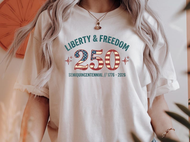 Puede incluir: Camiseta color crema con el texto "Liberty & Freedom 250" en verde azulado y rojo, celebrando el semiquincentenario. El n&uacute;mero 2 y el cero est&aacute;n dise&ntilde;ados con un patr&oacute;n de bandera patri&oacute;tica. La camiseta tambi&eacute;n incluye los a&ntilde;os 1776-2026.