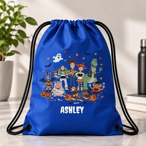 May include: A royal blue drawstring backpack featuring a colorful graphic of Toy Story characters dressed in Halloween costumes, including pumpkins, bats, and autumn leaves. The name ASHLEY is printed in white capital letters at the bottom center.