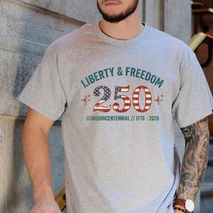 Puede incluir: Una camiseta gris con el texto "LIBERTY & FREEDOM 250 SEMIQUINCENTENNIAL // 1776-2026" en verde azulado y rojo. El n&uacute;mero 250 est&aacute; dise&ntilde;ado con un patr&oacute;n de estrellas y rayas. La camiseta es de manga corta.