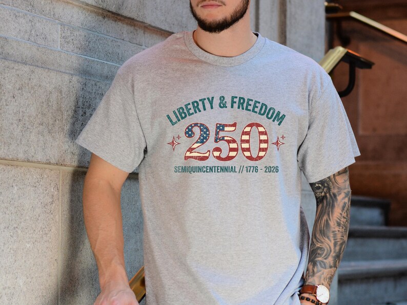 Puede incluir: Una camiseta gris con el texto "LIBERTY & FREEDOM 250 SEMIQUINCENTENNIAL // 1776-2026" en verde azulado y rojo. El n&uacute;mero 250 est&aacute; dise&ntilde;ado con un patr&oacute;n de estrellas y rayas. La camiseta es de manga corta.