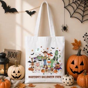 May include: A white canvas tote bag featuring a colorful graphic of Toy Story characters dressed in Halloween costumes, including a ghost, bats, and pumpkins. The text "MADISON'S HALLOWEEN CREW" is printed in brown letters at the bottom of the bag.