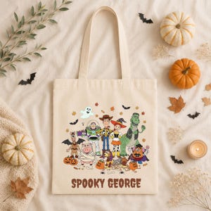 May include: A beige canvas tote bag featuring a colorful graphic of Toy Story characters dressed in Halloween costumes, including a mummy, a witch, and a vampire. The text SPOOKY GEORGE is printed in brown letters at the bottom of the bag.