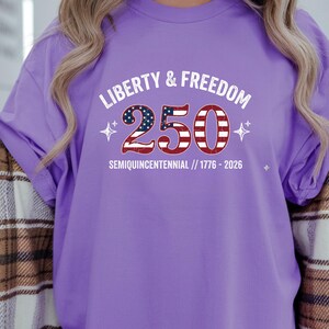 Puede incluir: Camiseta lila con la inscripci&oacute;n "LIBERTY & FREEDOM 250 SEMIQUICENTENNIAL // 1776-2026". El n&uacute;mero 250 est&aacute; dise&ntilde;ado con un patr&oacute;n de bandera americana roja, blanca y azul. La camiseta tiene mangas cortas.