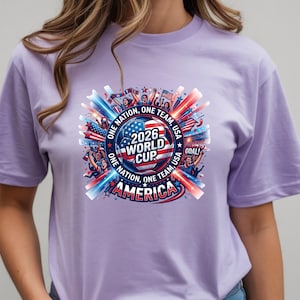 May include: A light purple t-shirt featuring a colorful graphic for the 2026 World Cup in America. The design includes a soccer ball, the American flag, and the text "ONE NATION, ONE TEAM, USA". The shirt has short sleeves.