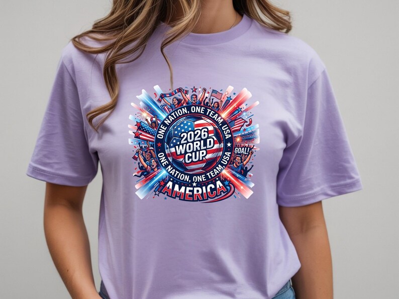 May include: A light purple t-shirt featuring a colorful graphic for the 2026 World Cup in America. The design includes a soccer ball, the American flag, and the text "ONE NATION, ONE TEAM, USA". The shirt has short sleeves.