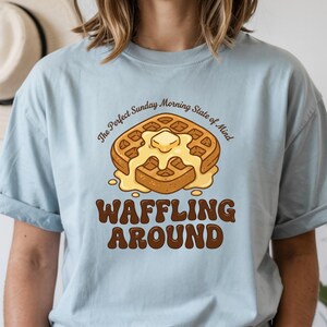 Może przedstawiać: Jasnoniebieska koszulka z grafiką gofra. Gofry mają masło i tekst "The Perfect Sunday Morning State of Mind" i "Waffling Around" w kolorze brązowym. Swobodny t-shirt dla miłośnik&oacute;w śniadań.
