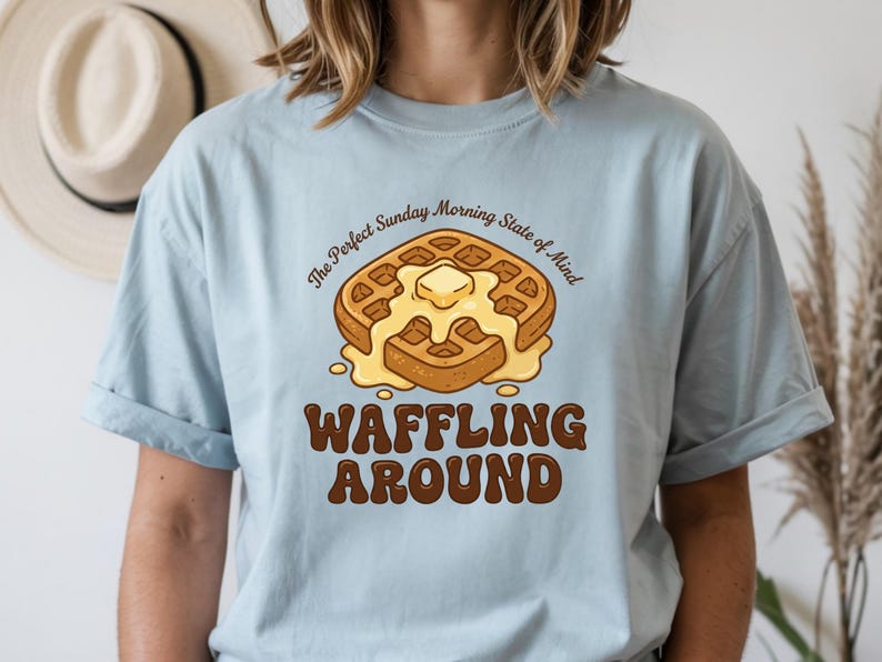 Może przedstawiać: Jasnoniebieska koszulka z grafiką gofra. Gofry mają masło i tekst "The Perfect Sunday Morning State of Mind" i "Waffling Around" w kolorze brązowym. Swobodny t-shirt dla miłośnik&oacute;w śniadań.