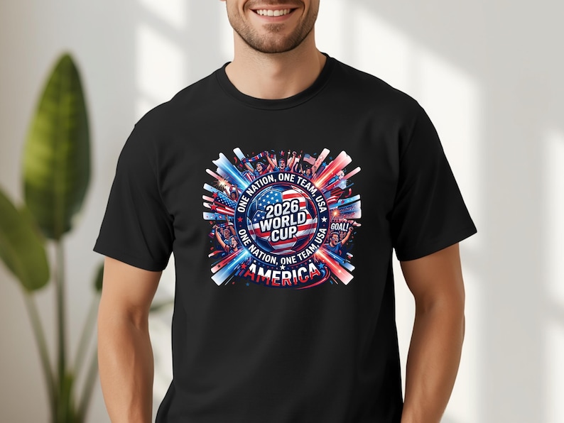 May include: Black t-shirt featuring a colorful graphic design for the 2026 World Cup in the USA. The design includes the text "ONE NATION, ONE TEAM, USA" and "AMERICA" with red, white, and blue accents.