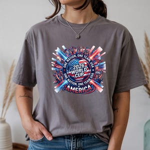 May include: A gray t-shirt featuring a colorful graphic for the 2026 World Cup in America. The design includes the text "2026 World Cup America" with red, white, and blue accents, and the words "One Nation, One Team, Goal!"